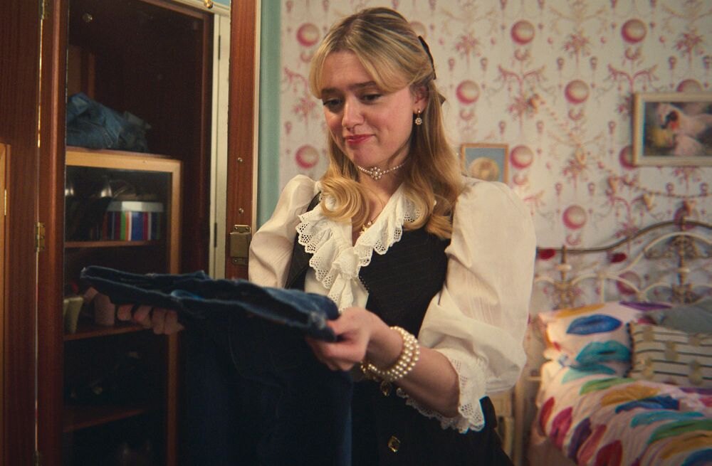 Aimee Lou Wood jako Aimee Gibbs v seriálu Sexuální výchova (4. řada) drží v ruce své džíny.