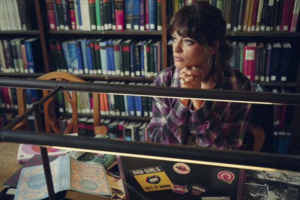 Emma Mackey jako Maeve Wiley v Sexuální výchově sedí u stolu a zamyšleně se dívá do strany.